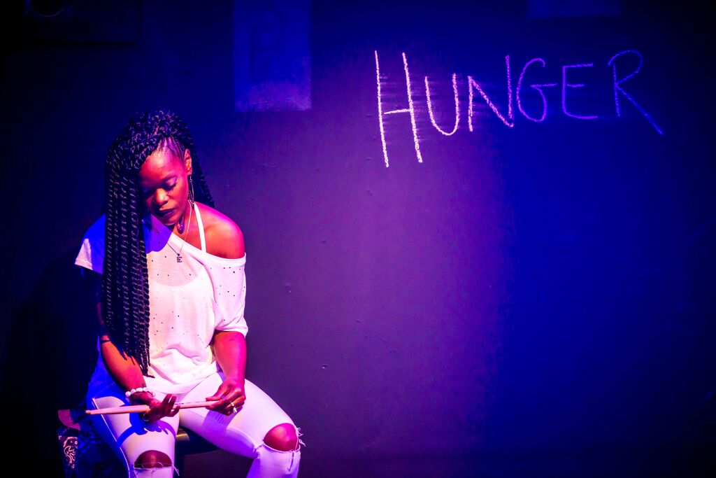 Photo of Ebony Stewart wearing all white. She is a dark skinned Black woman sitting down holding a pair of drumsticks. The lighting is a soft purple with the word Hunger scrawled in white chalk on the wall behind her.