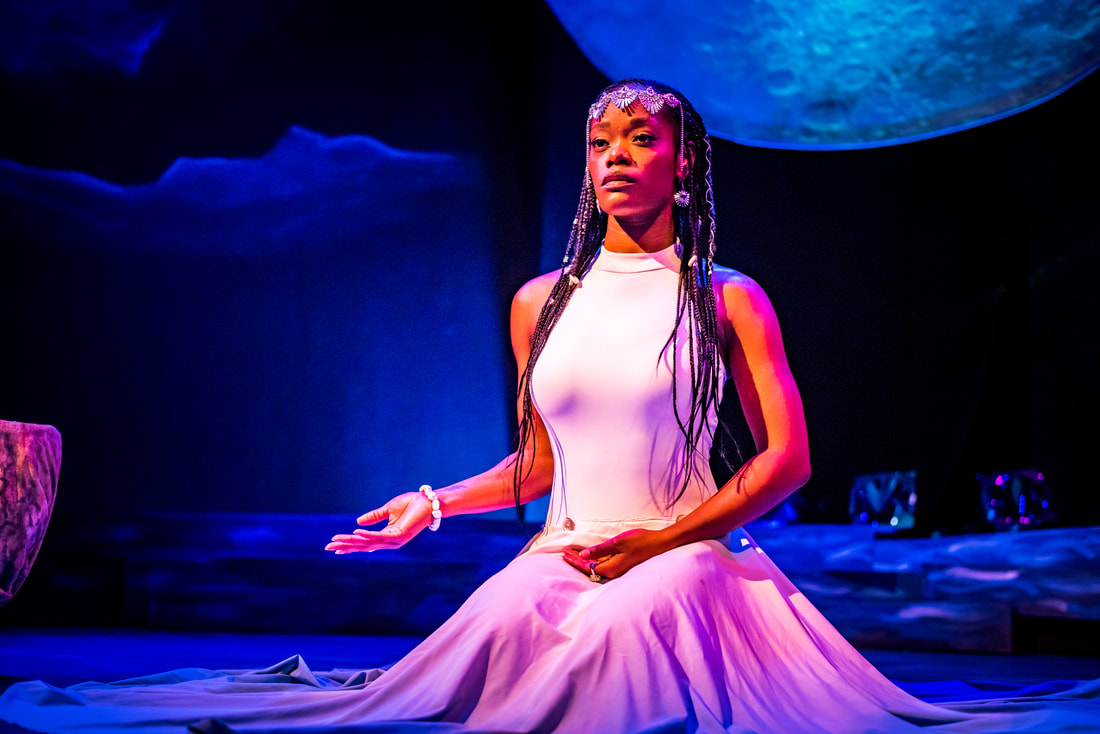 Ebony Stewart, a dark skinned Black woman, is pictured wearing a white dress that encircles her as she kneels on a theater floor. Her hands are folded serenely in her lap and she is lit by a soft purple glow. A projection of the moon appears on the dark wall behind her.