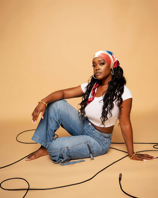 Ebony Stewart, a dark-skinned Black woman, stares regally at the viewer. She is wearing a large spiky gold crown with flowers at the base, large oval earrings, a thick gold necklace, and a white t shirt that says Black women don't miss.